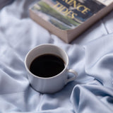 White mug with coffee on a soft blue fabric surface, with a book titled 'Prince Tides' partially visible.